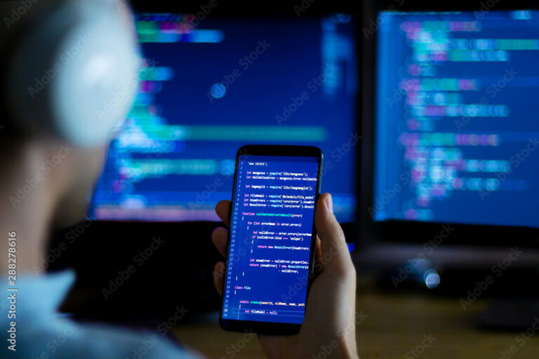 Software developer freelancer with white headphones and glasses working with program code: C++ Java, Javascript on wide displays and smartphone at night. Develops new web, desktop, mobile application.